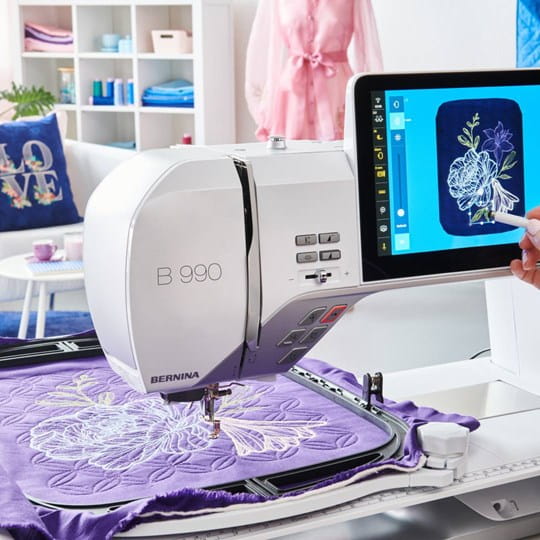 Bernina 990 machine à coudre avec grand écran tactile et broderie automatisée, réalisant un motif floral précis sur un tissu violet.