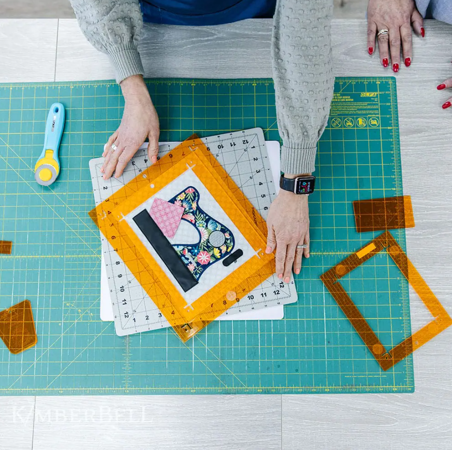 Vue de dessus de deux personnes utilisant une règle Orange Pop rectangulaire pour positionner et découper un appliqué de machine à coudre. Plusieurs cadres Orange Pop et un cutter rotatif sont visibles sur la table de travail.