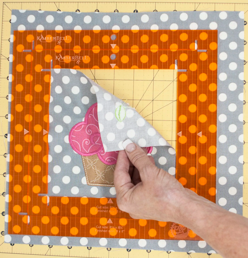 La règle Orange Pop de Kimberbell encadre un appliqué cupcake sur un tissu, facilitant le positionnement et la découpe de motifs complexes.