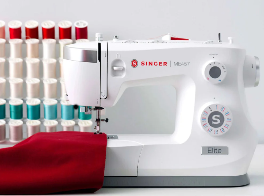 Machine à coudre SINGER Elite ME457 – Machine en cours d’utilisation, avec un tissu rouge sous l’aiguille, et un présentoir de bobines en arrière-plan.