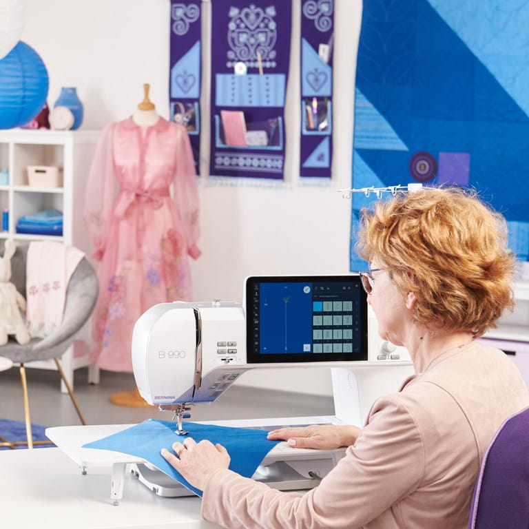 Utilisation de la Bernina 990 machine à coudre – Femme cousant un tissu bleu sur la machine dans un atelier de couture décoré de patchworks et vêtements.