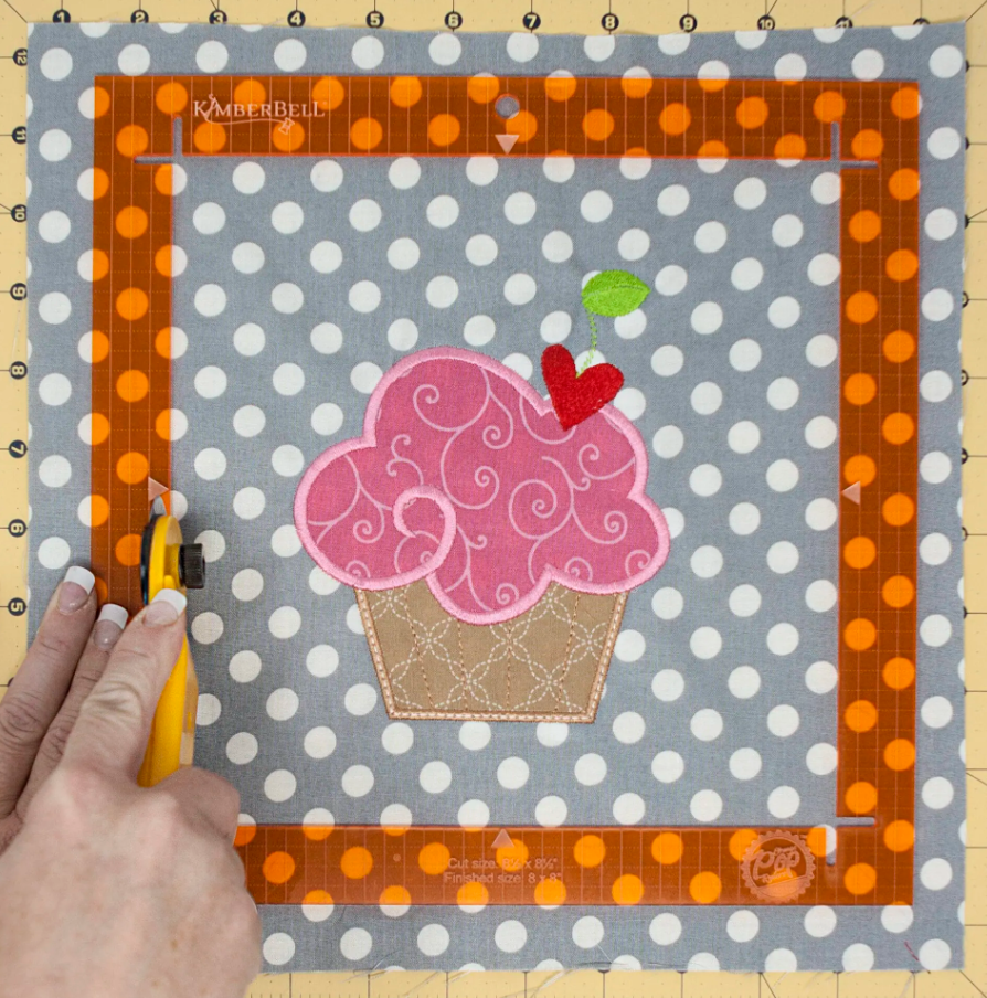 Utilisation de la règle Orange Pop sur un tissu à pois gris avec un appliqué cupcake rose. La règle est positionnée pour couper précisément autour du motif à l'aide d'un cutter rotatif.