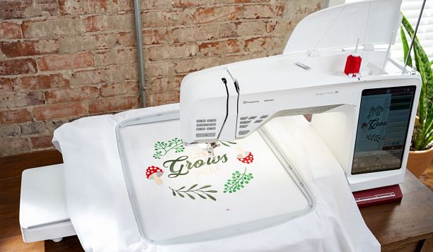 Machine à broder Husqvarna Viking DESIGNER RUBY™ 90 en action – Broderie d’un motif floral avec champignons et texte "Grows" sur un tissu blanc, illustrant la précision et la finesse des détails réalisés par la machine.