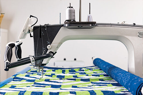Machine à quilter Grace Q'nique 19X Elite avec une structure robuste, poignées ergonomiques et bobine de fil argenté, en train de piquer un quilt aux motifs verts et bleus.