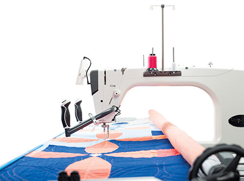 Machine à quilter Q'nique 21X Elite, équipée d'un bras allongé et d'un design ergonomique, avec une bobine de fil rose, utilisée pour piquer un quilt aux motifs bleus et oranges.