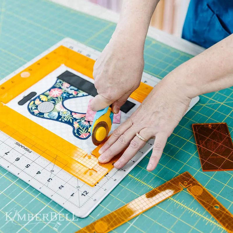 Gros plan sur une personne utilisant une règle Orange Pop rectangulaire et un cutter rotatif pour découper précisément un appliqué de machine à coudre coloré sur du tissu.