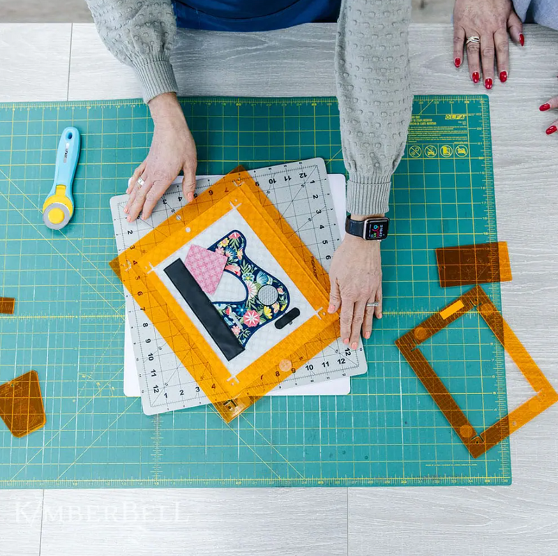  Vue de dessus de deux personnes utilisant une règle Orange Pop rectangulaire pour positionner et découper un appliqué de machine à coudre. Plusieurs cadres Orange Pop et un cutter rotatif sont visibles sur la table de travail.