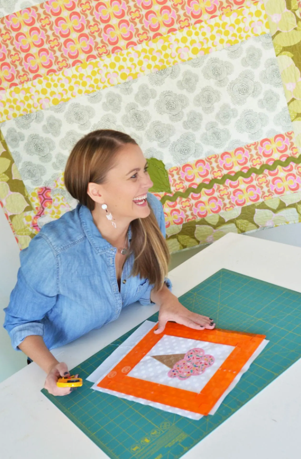 Femme souriante portant une chemise en jean, travaillant sur un appliqué encadré par une règle Orange Pop de Kimberbell sur une table de couture. Elle est prête à découper son bloc avec un cutter rotatif.