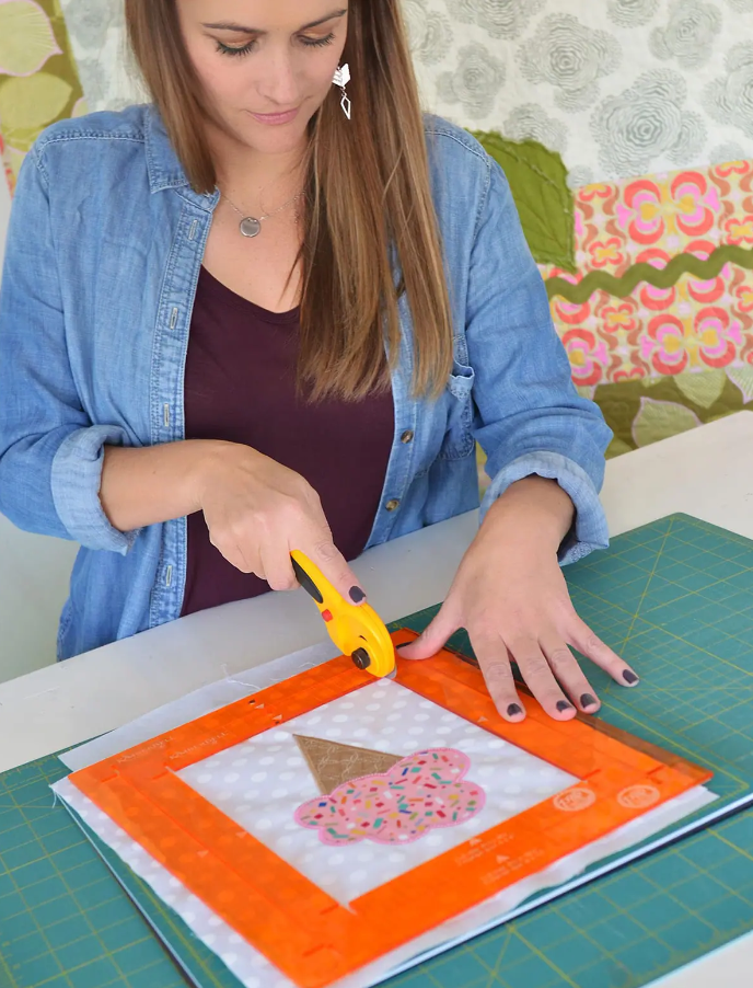 Gros plan d’une femme utilisant un cutter rotatif sur une règle Orange Pop pour découper un appliqué cupcake sur un tissu à pois.